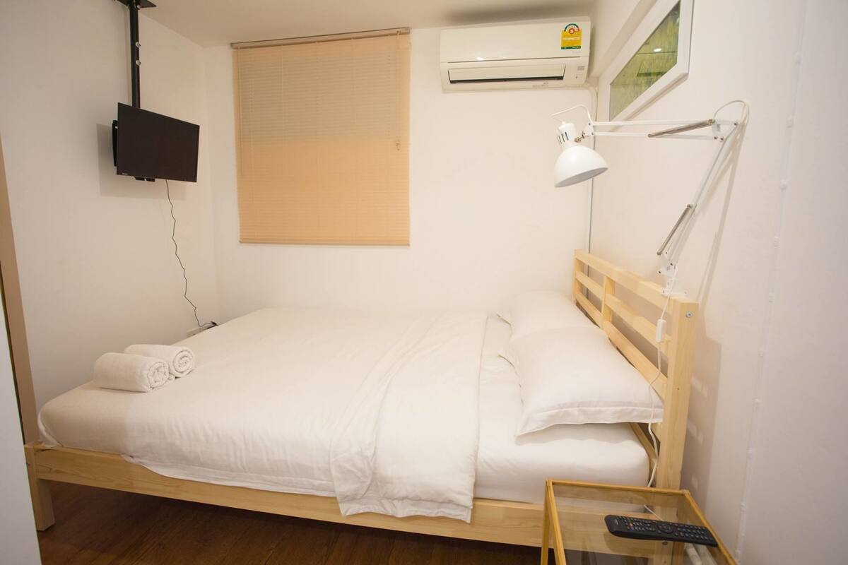 Un lit simple avec un cadre en bois, une literie blanche, une lampe de chevet, un téléviseur fixé au mur et un appareil de climatisation.