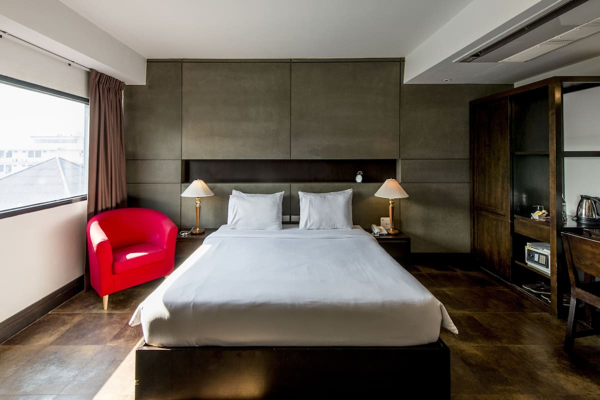A hotel room with a large bed, two bedside lamps, a red chair, a wooden wardrobe, and a view of the city through a window.