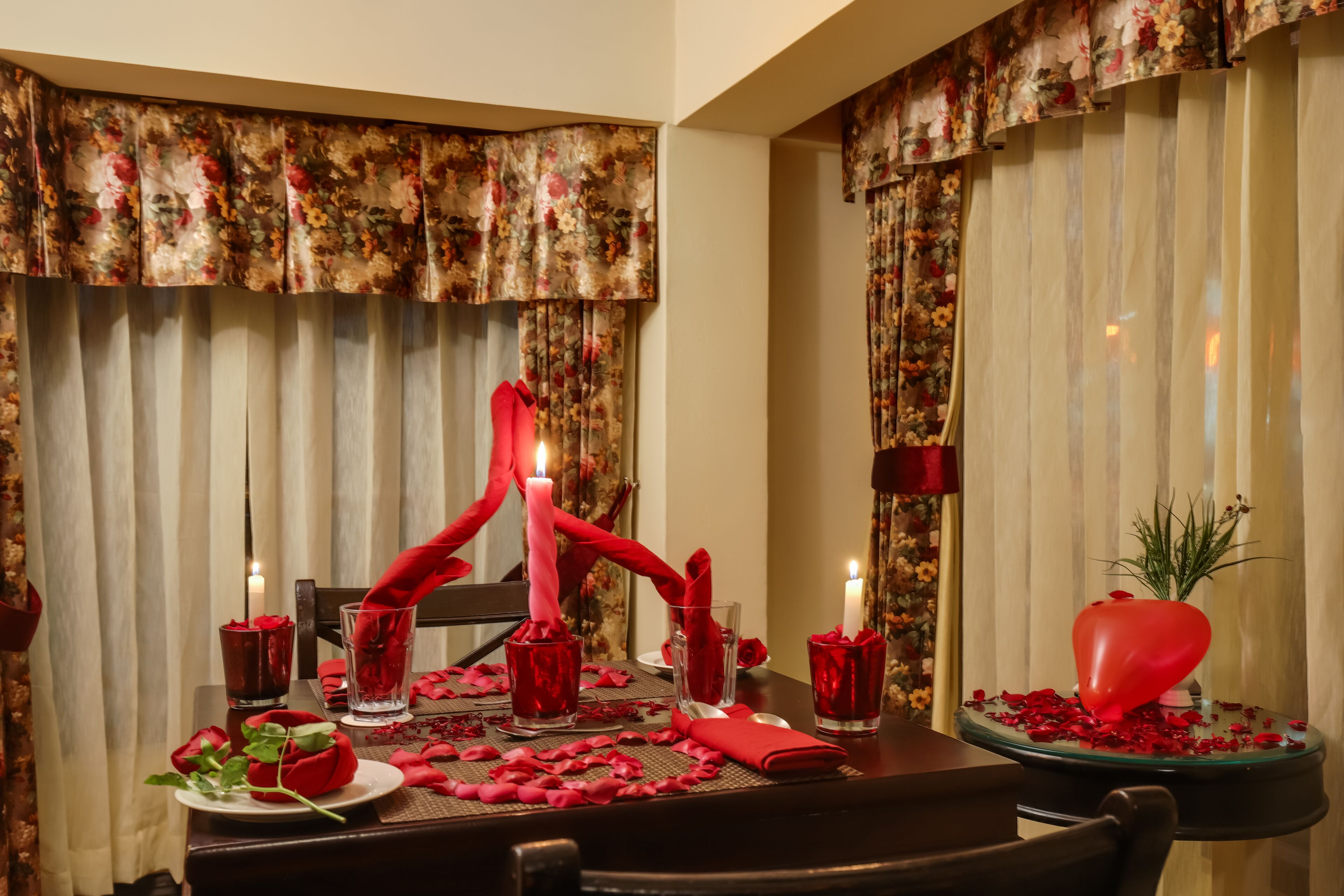 Ein romantisches Dinnerambiente mit floralen Vorhängen, Kerzen, Rosenblättern und einer herzförmigen Dekoration auf dem Tisch.