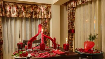 Ein romantisches Dinnerambiente mit floralen Vorhängen, Kerzen, Rosenblättern und einer herzförmigen Dekoration auf dem Tisch.