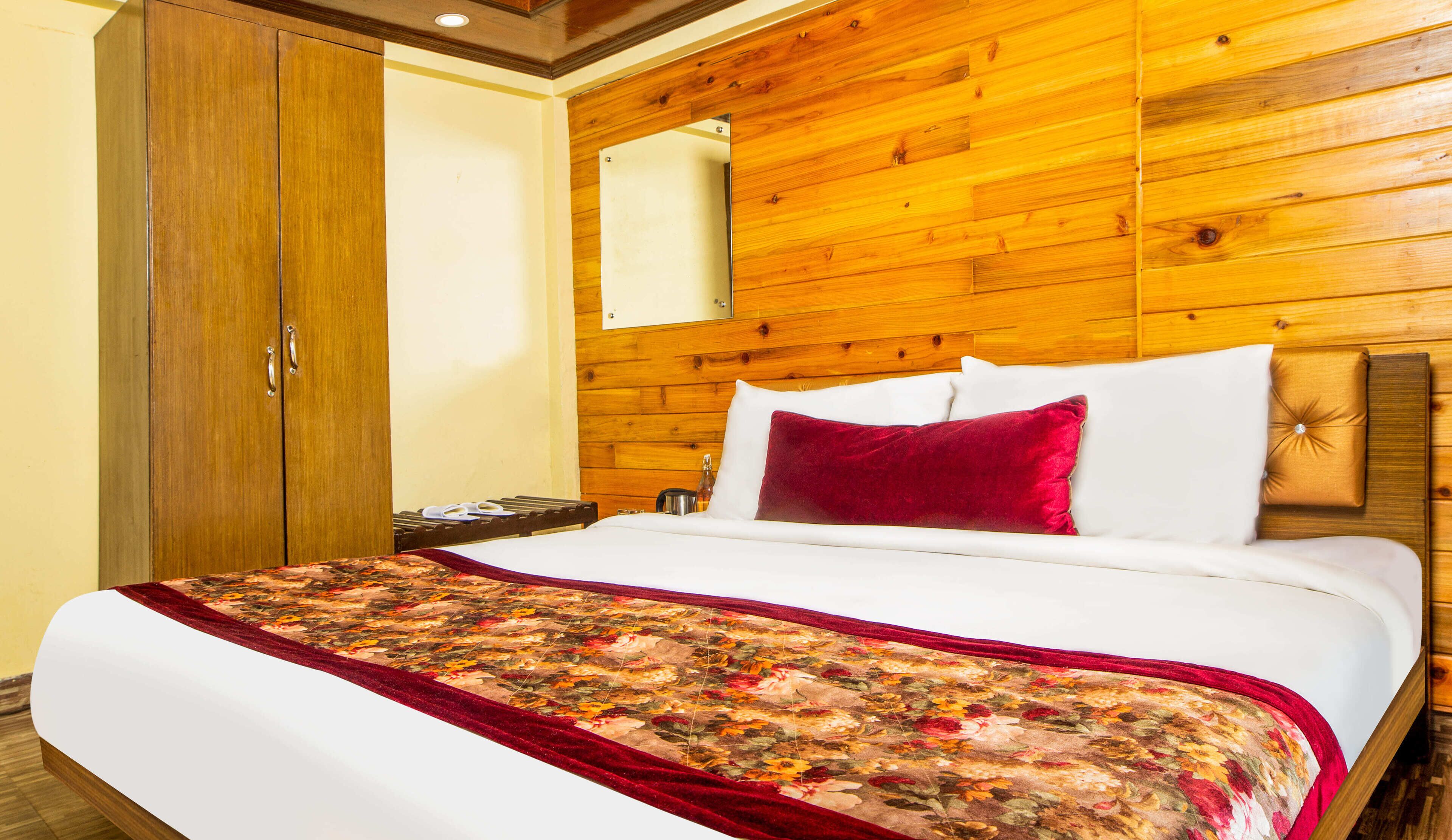 A hotel room with a wooden headboard, a large bed with white linens and a floral bedspread, a wooden wardrobe, and a mirror on the wall.