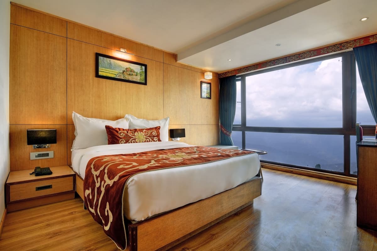 A hotel room with a large bed, wooden paneling, a nightstand with a phone, and a view of the sea through large windows.