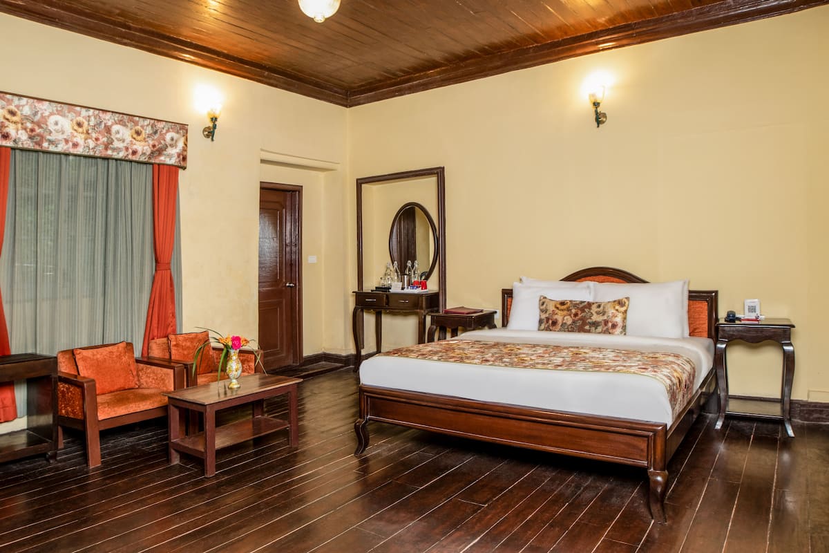 A hotel room with a wooden floor, a bed with a floral bedspread, a wooden nightstand, a desk with a chair, and a window with curtains.