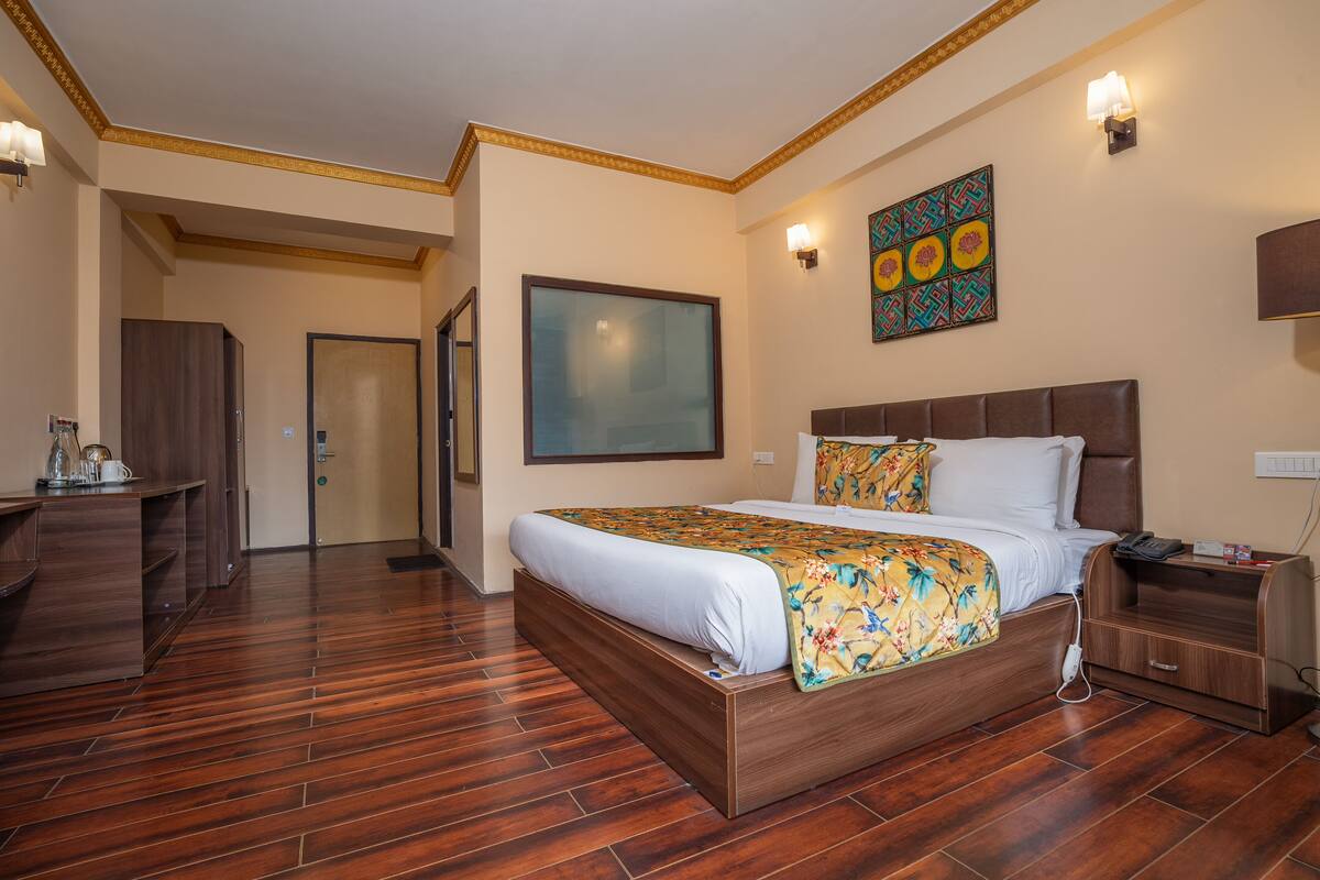 A hotel room with a wooden floor, a large bed with a floral bedspread, a nightstand with a phone, and a wall-mounted artwork.