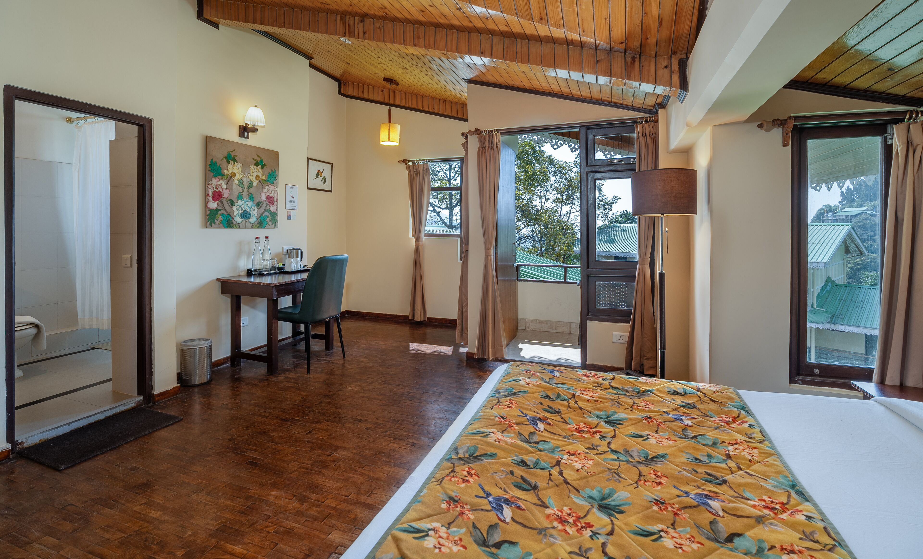 Habitación de hotel con techo de madera, cama con cubrecama floral, escritorio con silla, ventana con cortinas y vistas al exterior.