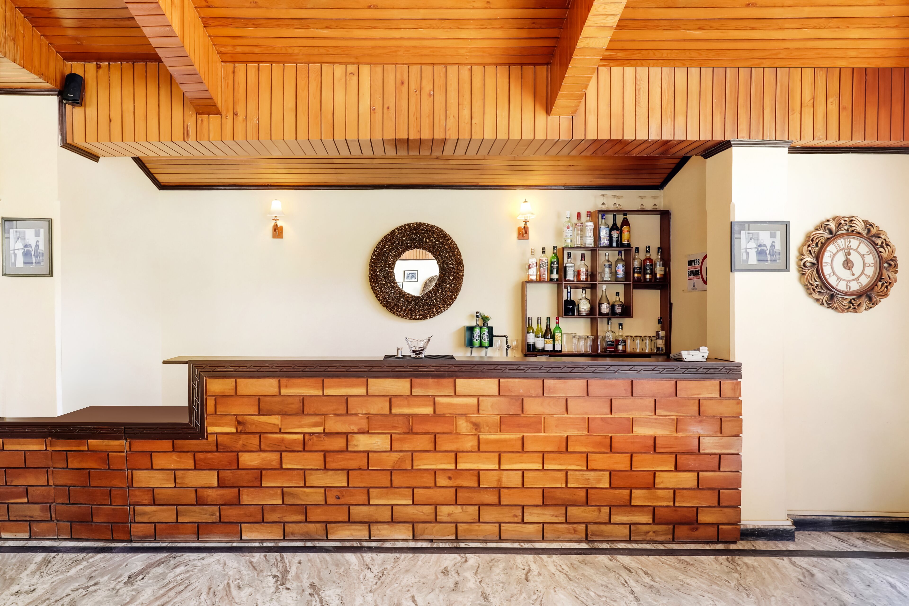 Un área de recepción con un mostrador de ladrillo, techo de madera, luces fijadas en la pared, un espejo, un reloj y un estante con botellas.