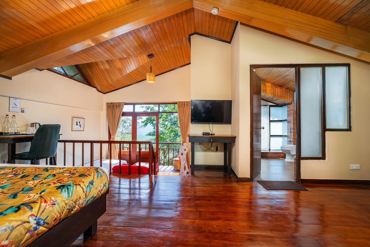 A spacious room with a wooden ceiling, a bed with a floral bedspread, a desk with a television, and a view of the outdoors.