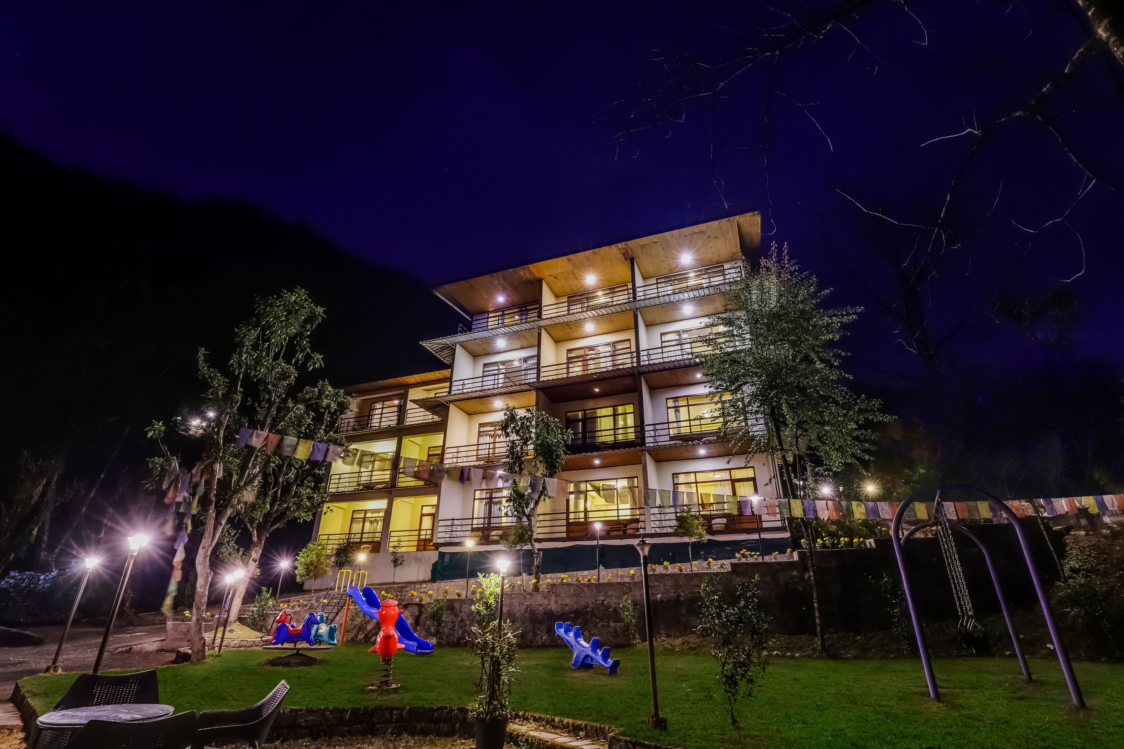 Un hotel de varios pisos con balcones, una zona de juegos para niños y un jardín iluminado durante la noche.
