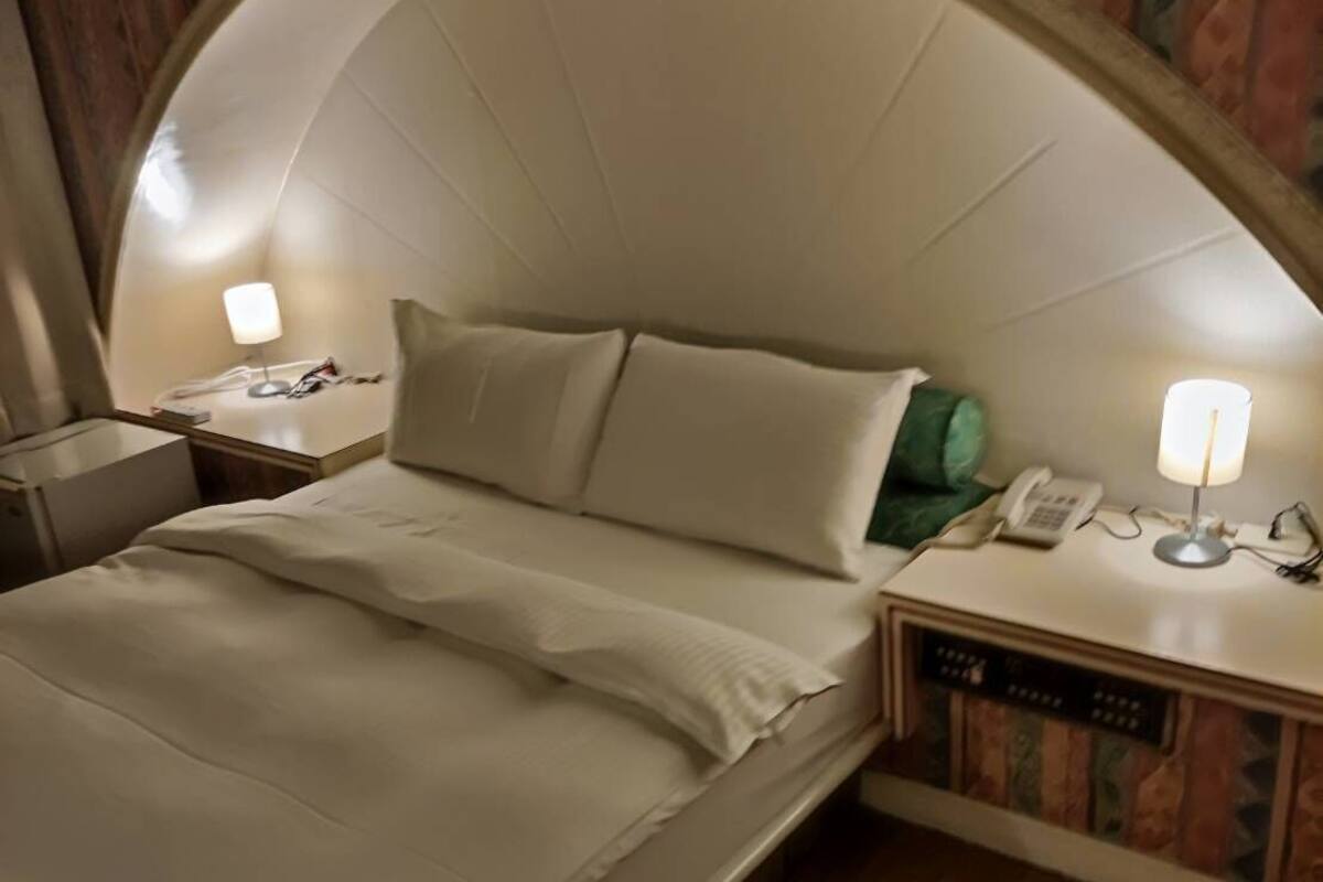 A hotel room with a curved headboard, a bed with white linens, two bedside tables with lamps, and a small bedside phone.
