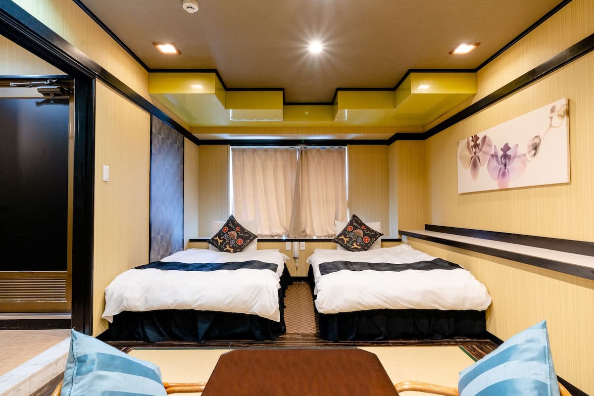 A hotel room with two beds, a wooden floor, a large window with curtains, and a painting on the wall.