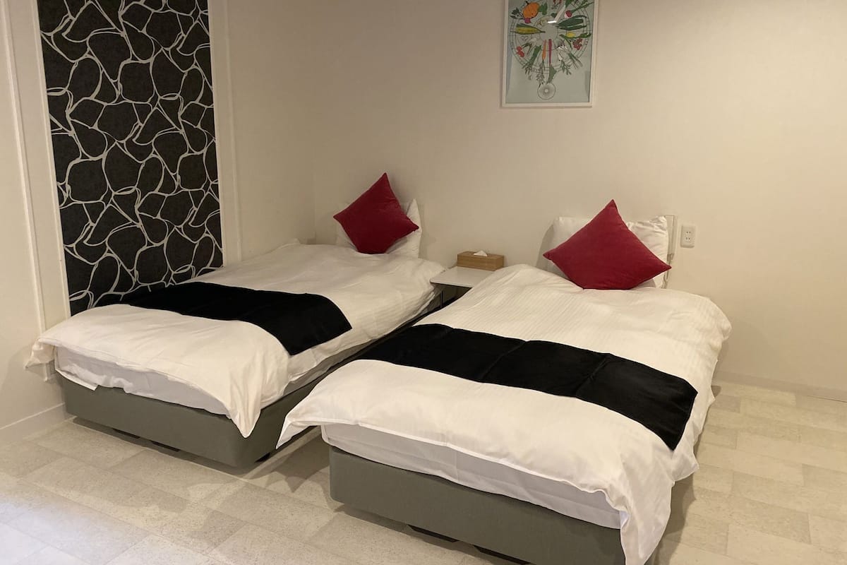 A room with two beds, each with white bedding and red pillows, a small table with a box, and a wall clock.