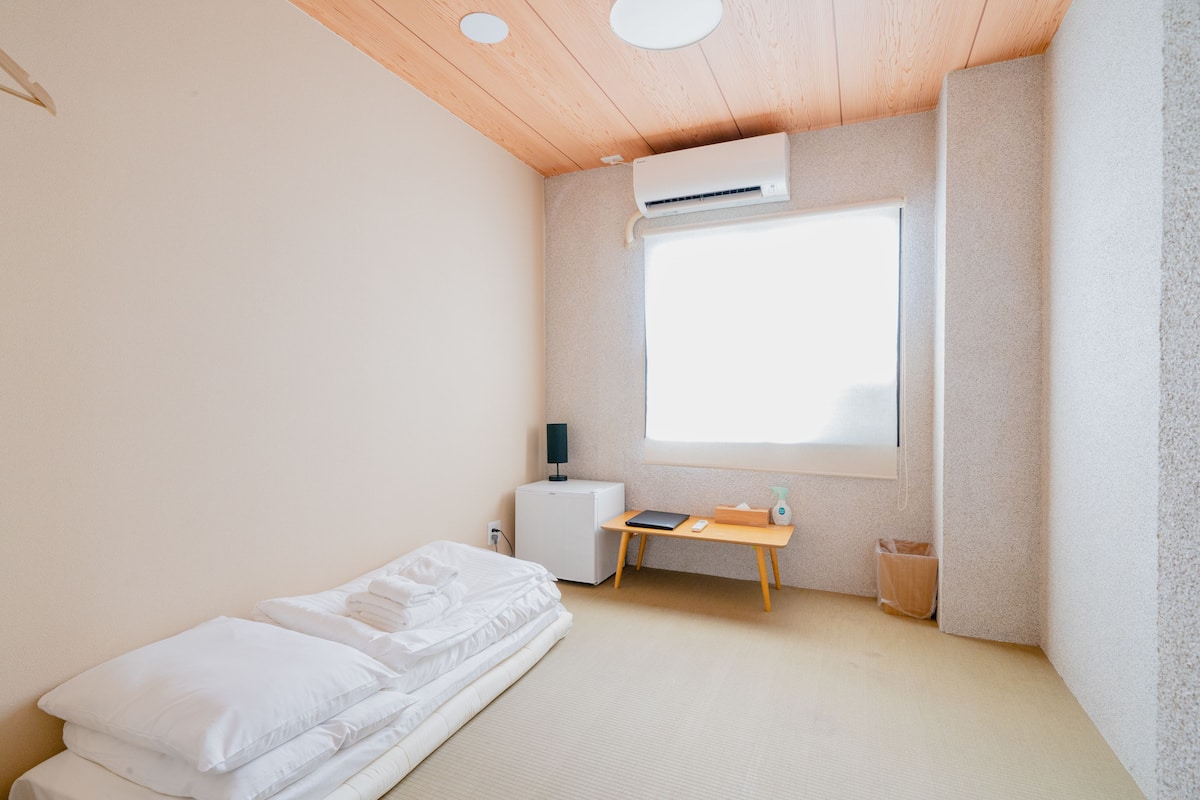 A minimalist room with a bed, a small desk, a window, and an air conditioning unit.