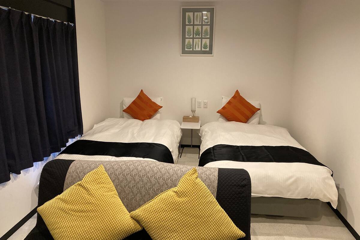 A hotel room with two beds, each with a white bedspread and orange pillows. There is a small table with a lamp and a framed picture on the wall.