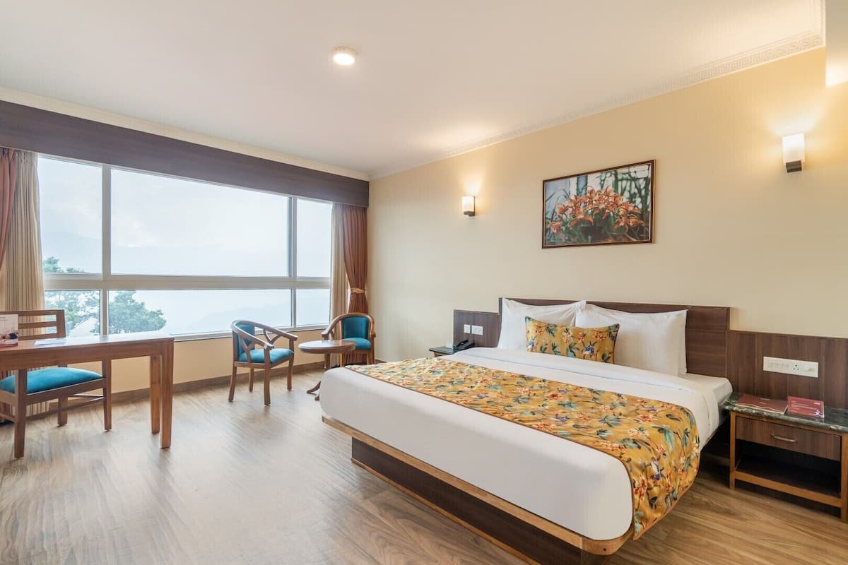 A hotel room with a large bed, a desk with chairs, a painting on the wall, and a window with curtains.
