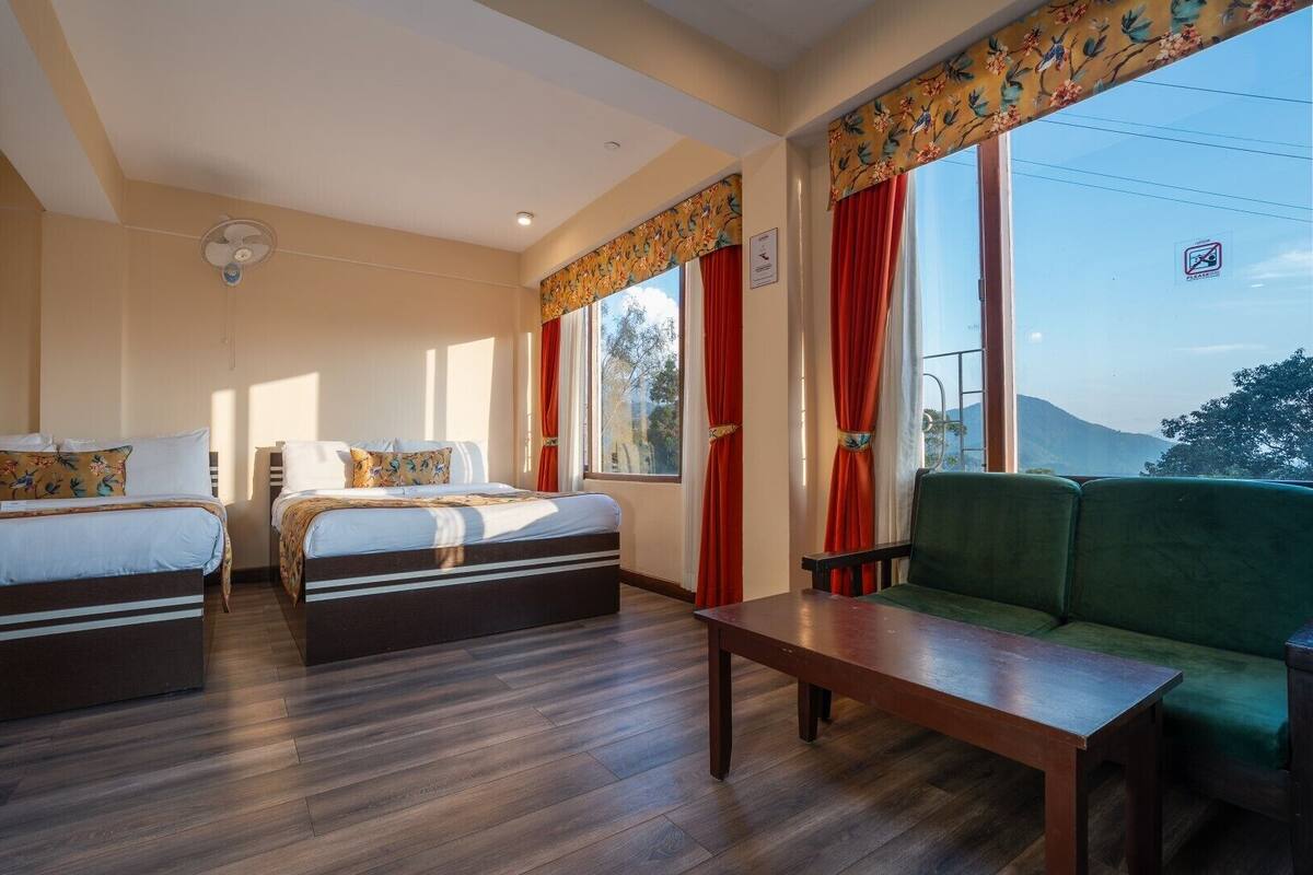 A hotel room with two beds, a green sofa, a wooden table, and large windows with red curtains.