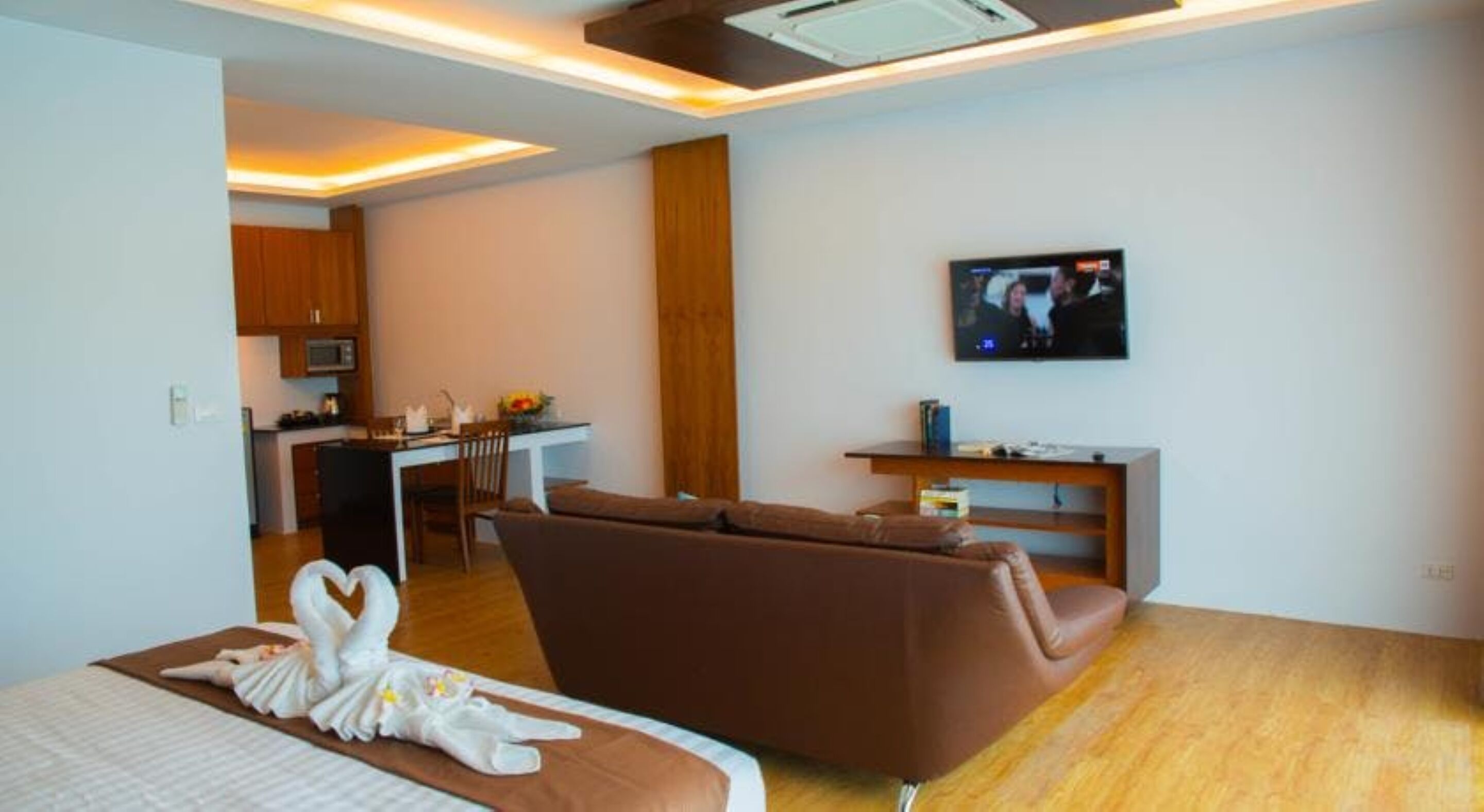 Una sala de estar moderna con un sofá marrón, un televisor de pantalla plana montado en la pared y una pequeña zona de cocina al fondo.