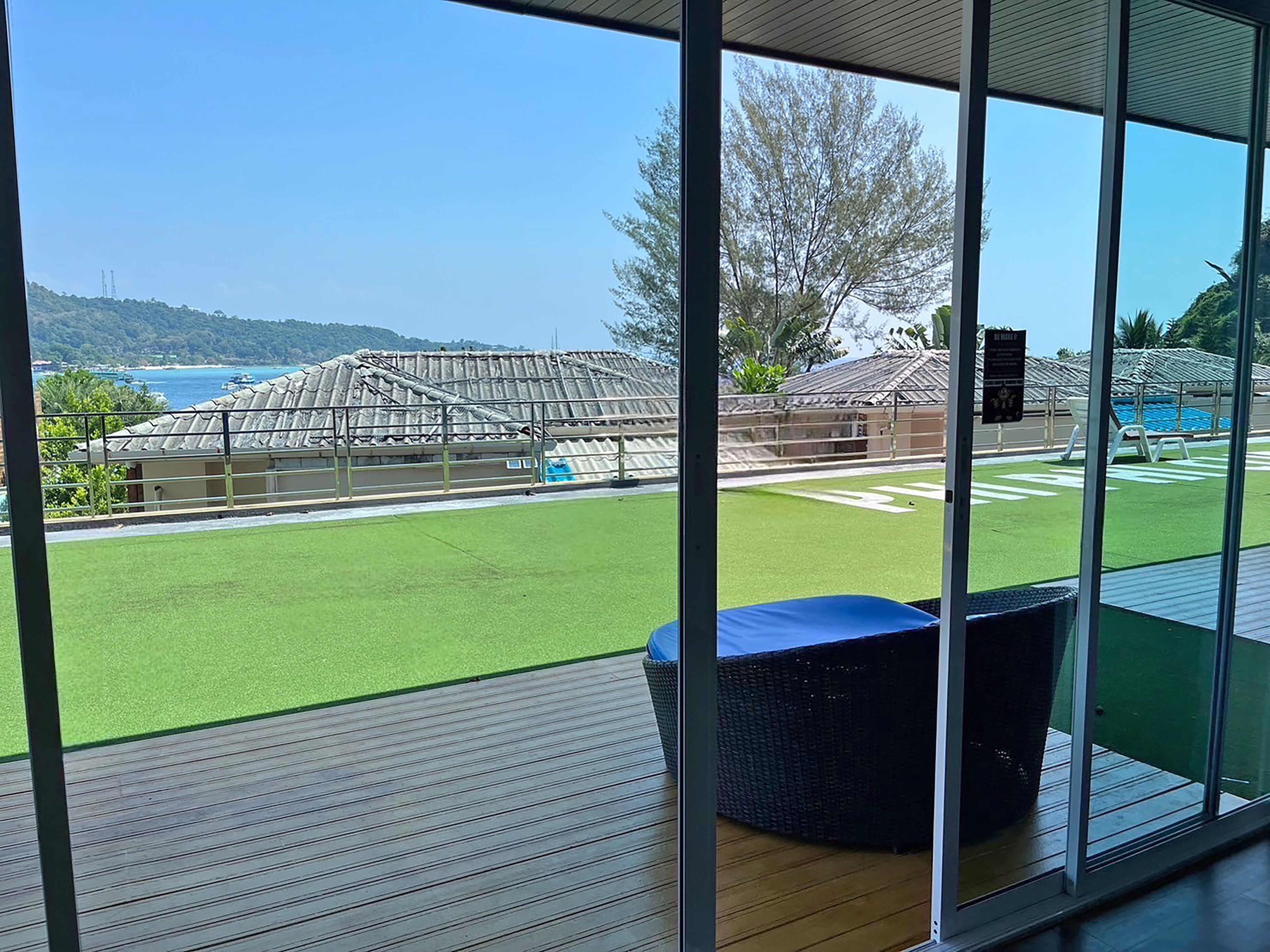 Vista a través de amplios ventanales de un área de piscina con césped artificial, una estructura cubierta y un cielo azul claro.
