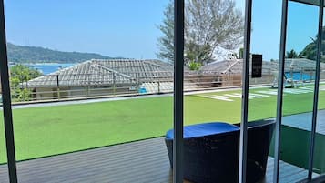 Vista a través de amplios ventanales de un área de piscina con césped artificial, una estructura cubierta y un cielo azul claro.