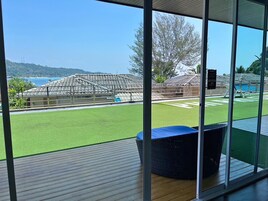 Vista a través de amplios ventanales de un área de piscina con césped artificial, una estructura cubierta y un cielo azul claro.