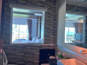 A hotel room with a large window, a bed, a bathroom with a mirror, and a small table with flowers.