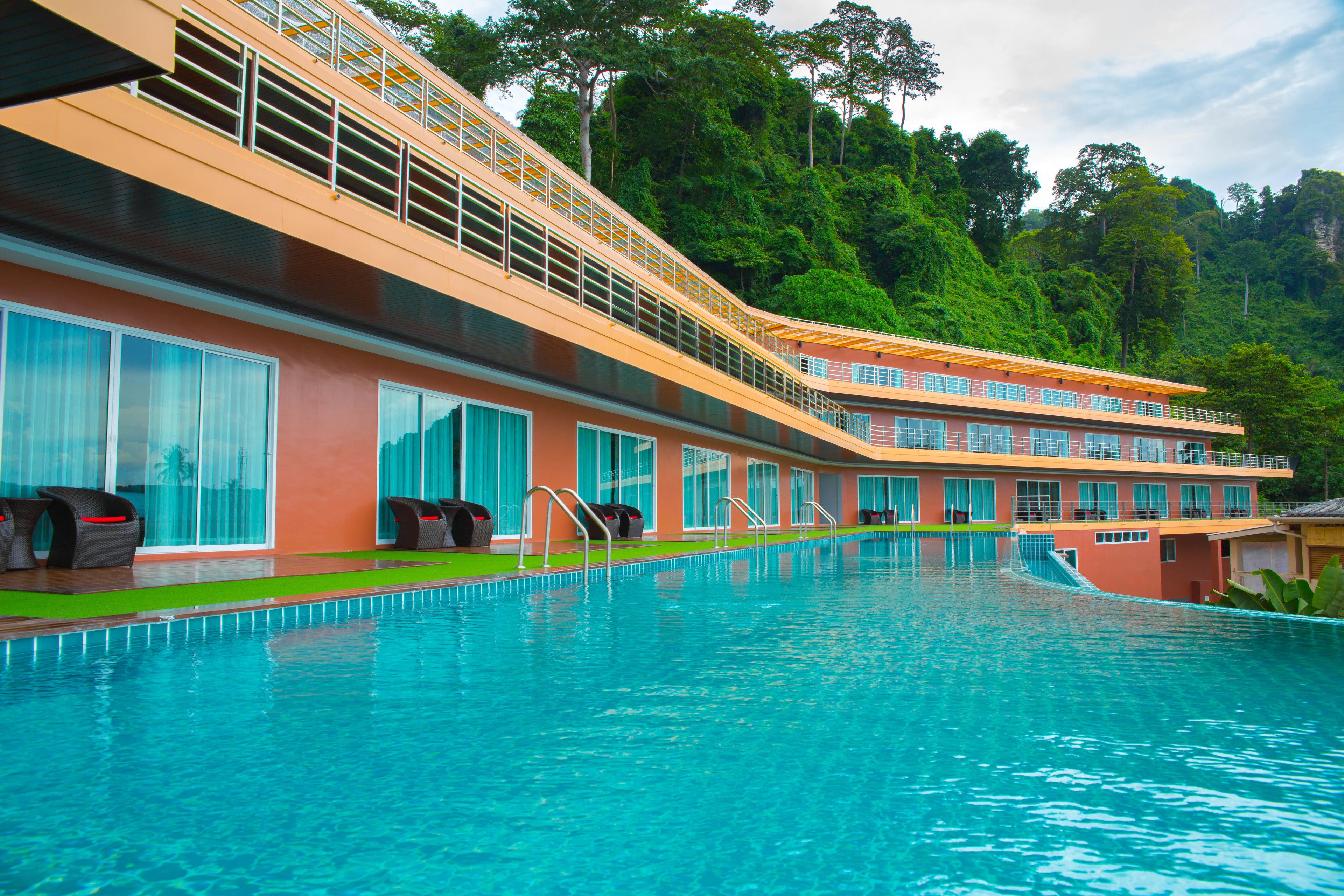 Et hotel med swimmingpool, flere balkoner og frodig, grøn skov i baggrunden.