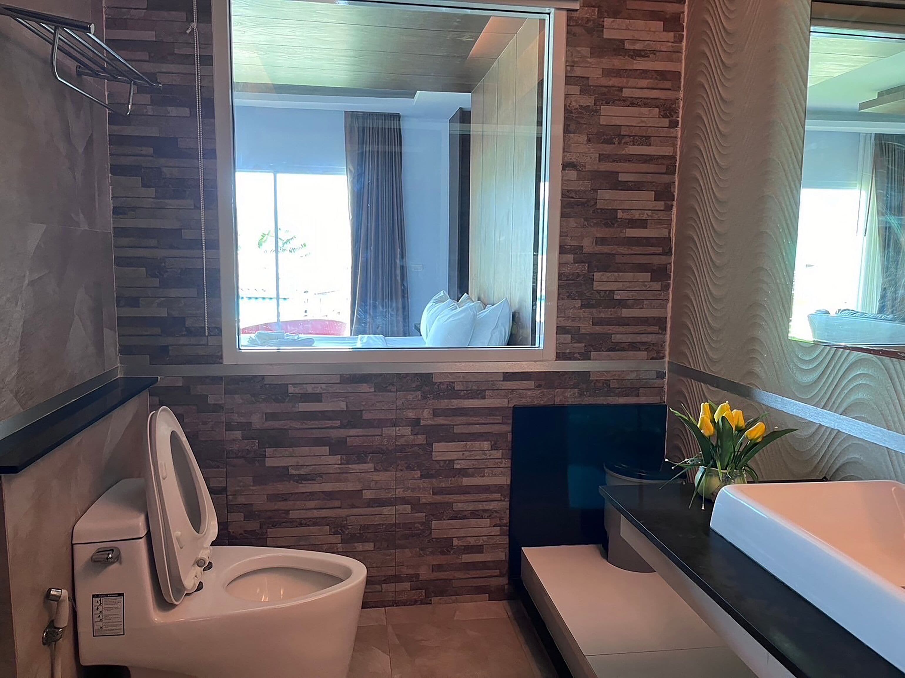A modern bathroom with a toilet, sink, and window overlooking a room with a bed and curtains.