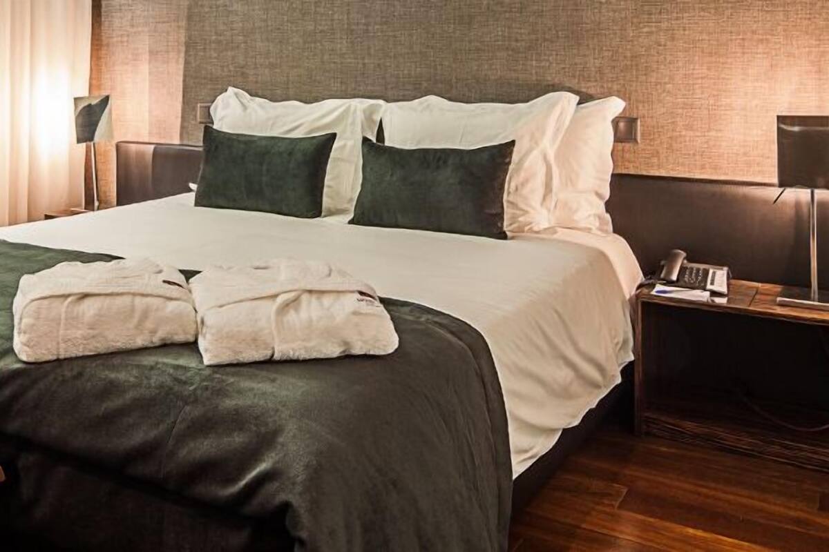 A neatly made bed with a green and white duvet, two pillows, and a bedside table with a lamp and a phone.