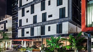 A multi-story hotel with a modern design, featuring a mix of glass and concrete facades, outdoor seating area with plants, and a visible restaurant or cafe.