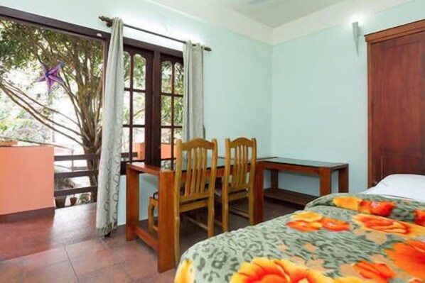 A room with a bed, a desk, and two chairs. There is a window with curtains and a view of trees outside.