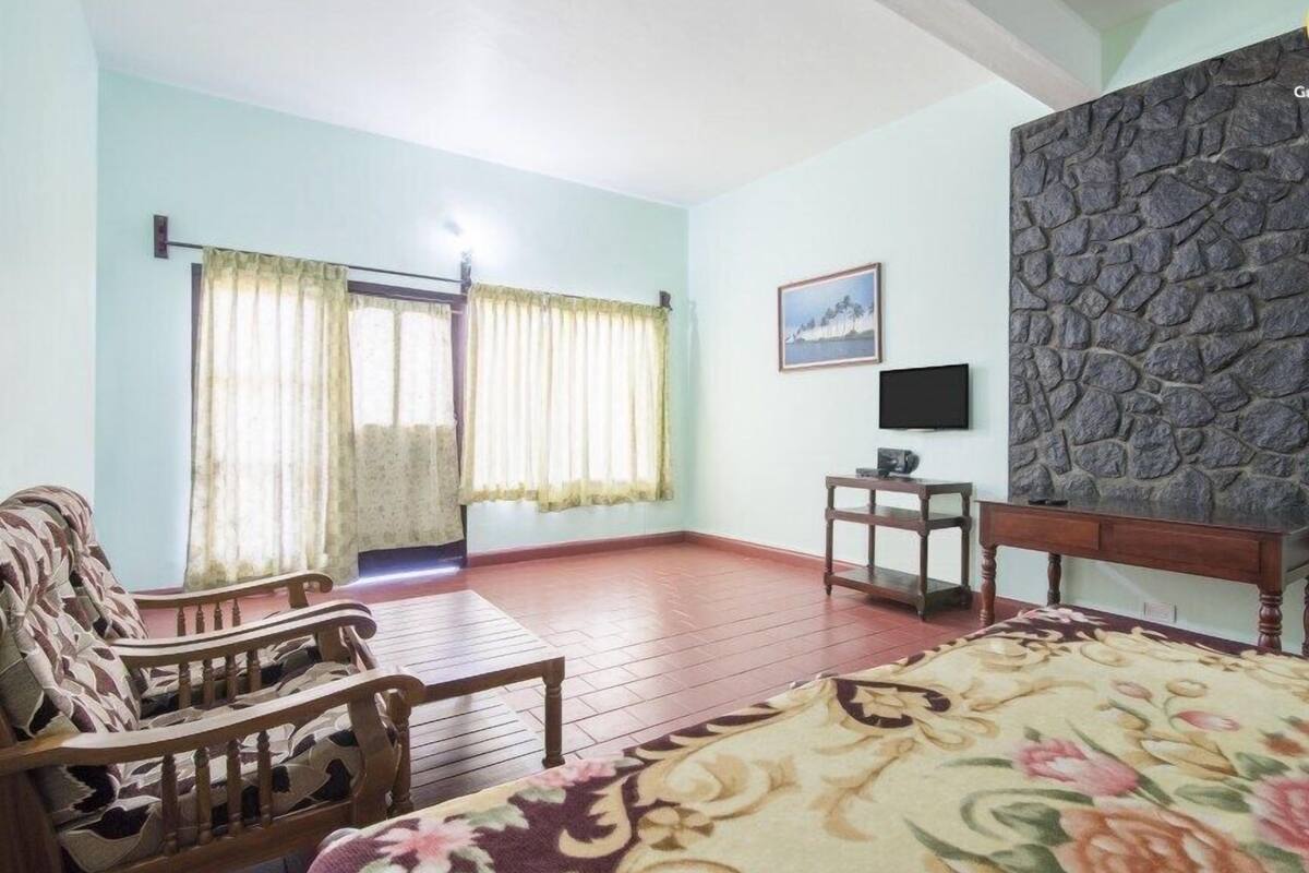 A room with a bed, a wooden chair, a small table, a TV, and a stone wall.