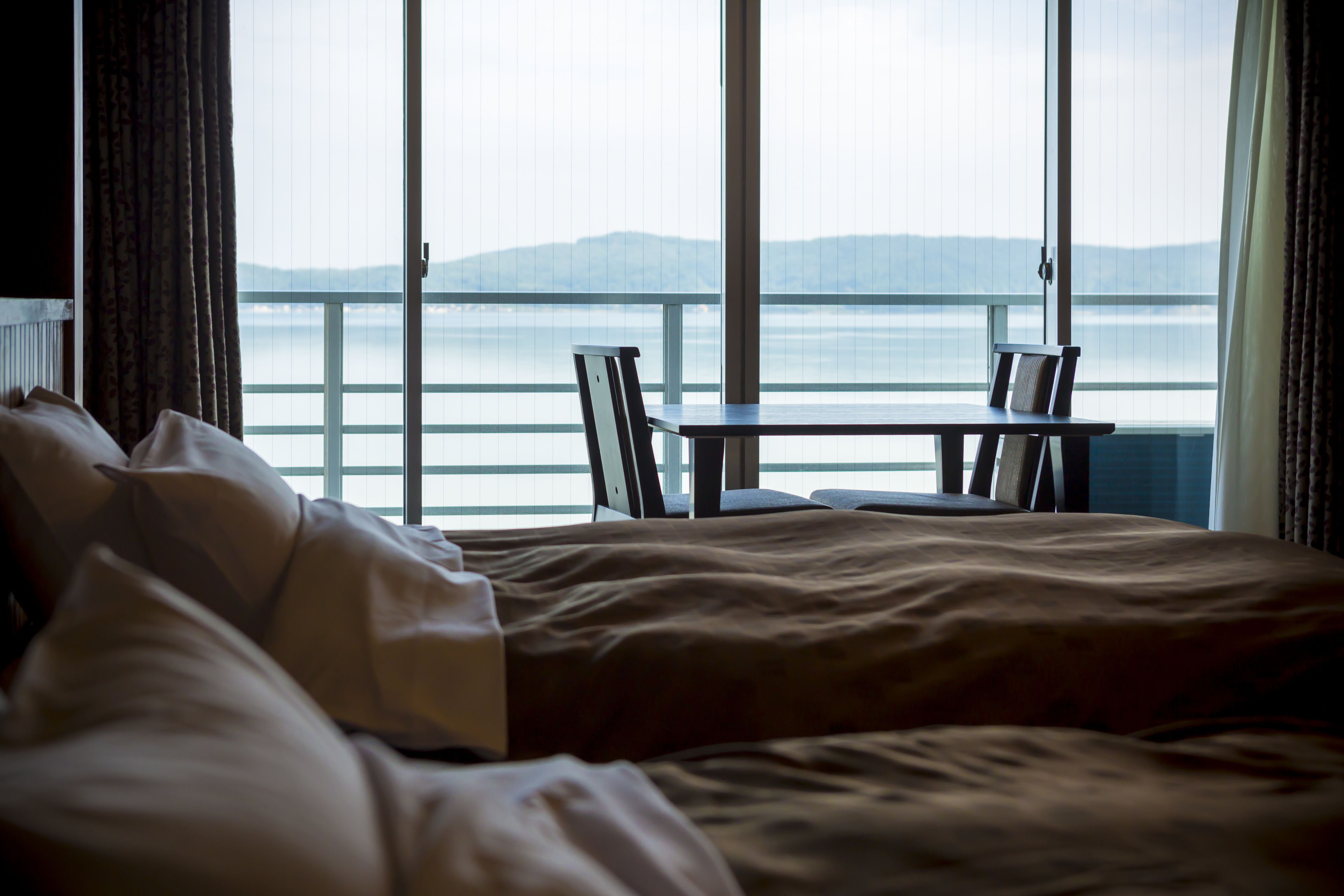 A hotel room with a bed, a small table, and chairs. The room has large windows with a view of the sea and mountains.