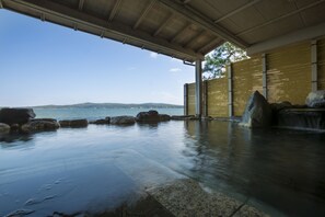 A hot spring area with a view of the sea and mountains, featuring large rocks and a wooden fence.