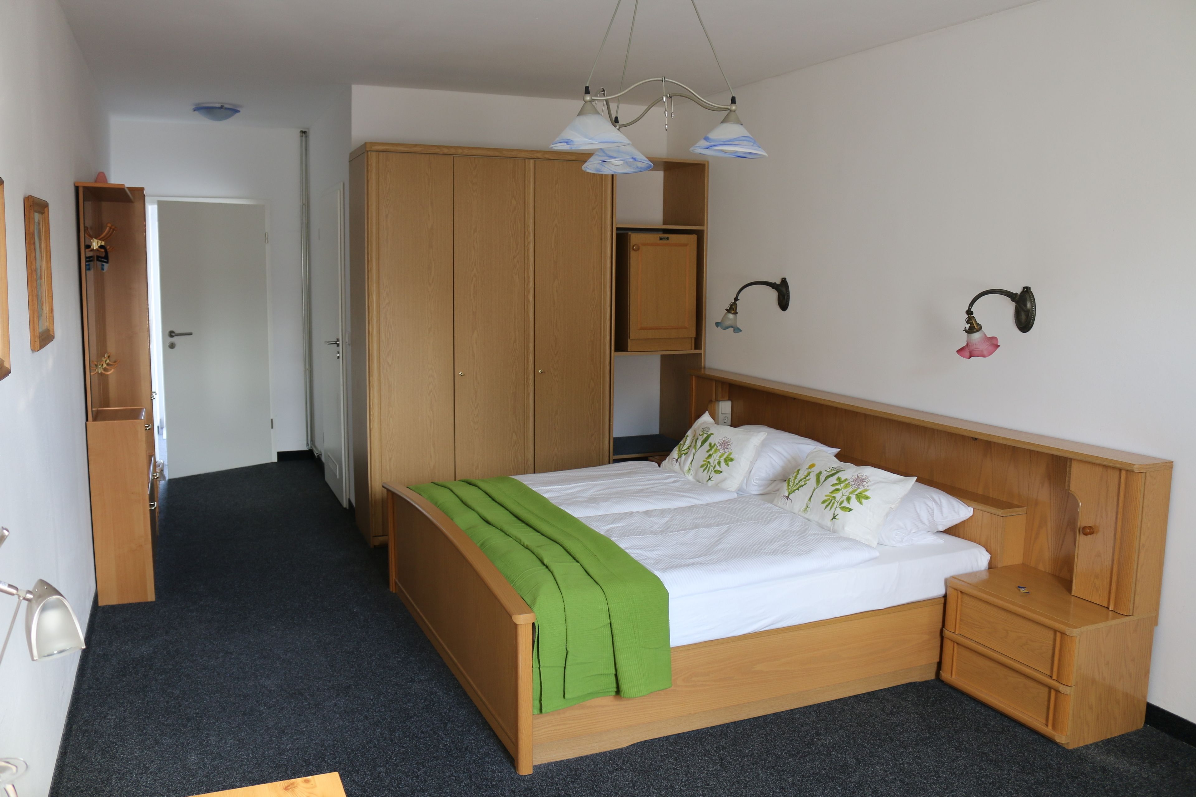 A bedroom with a wooden bed, a green throw blanket, a nightstand, a wardrobe, and a wall-mounted lamp.