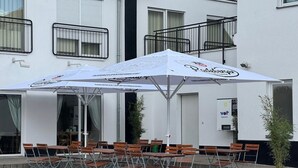 Un espace extérieur aménagé avec des tables et des chaises, un parasol blanc, et un bâtiment doté de balcons et de fenêtres.