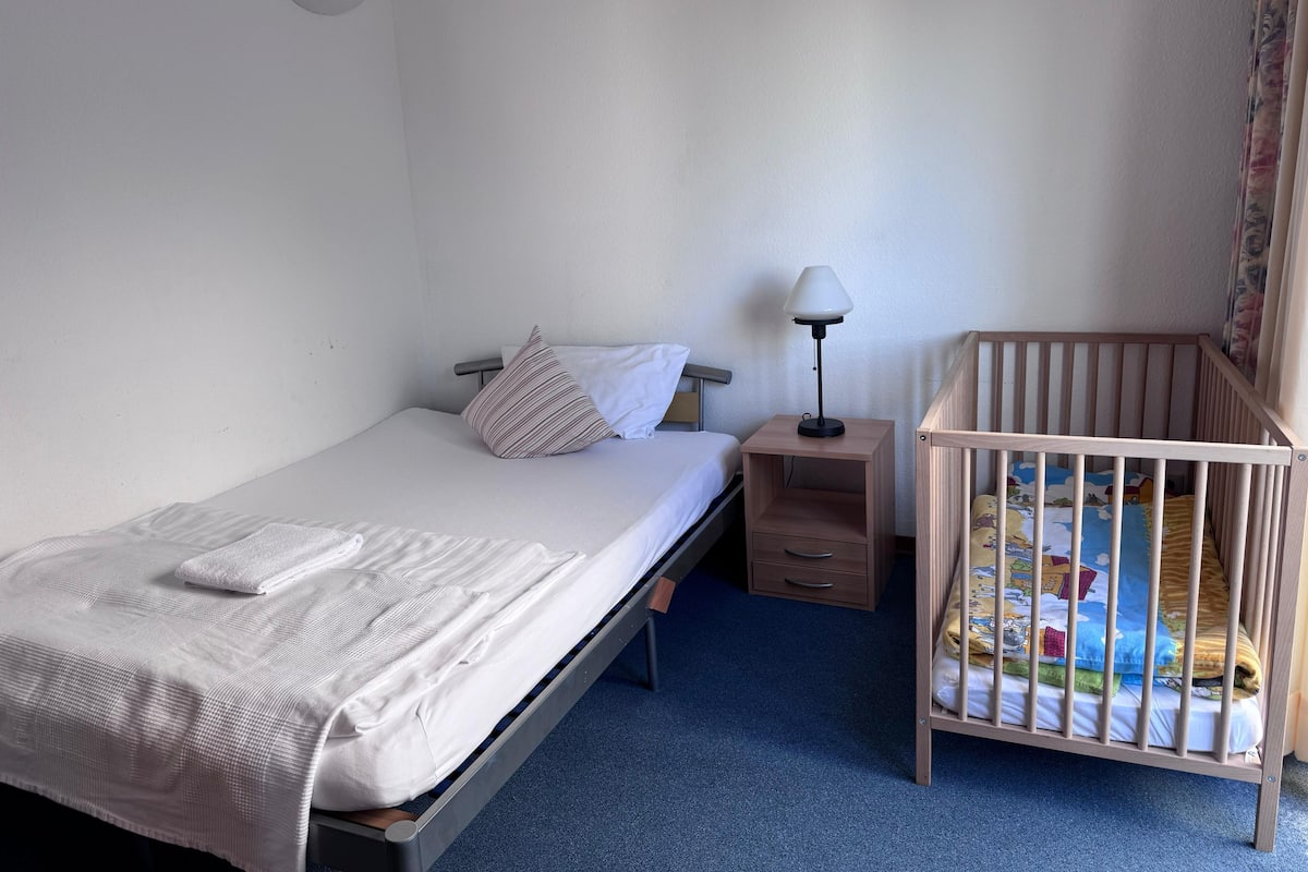 A single bed with a white mattress and a striped pillow, a bedside table with a lamp, a wooden crib with a colorful mattress, and a blue carpet.