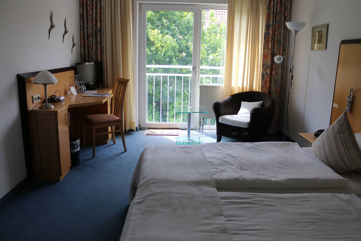 A hotel room with a bed, a desk with a computer, a chair, a television, and a balcony with a view of trees.