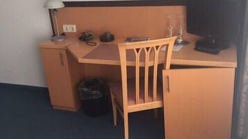 A compact hotel room with a wooden desk, a chair, a small trash bin, a lamp, and a television.