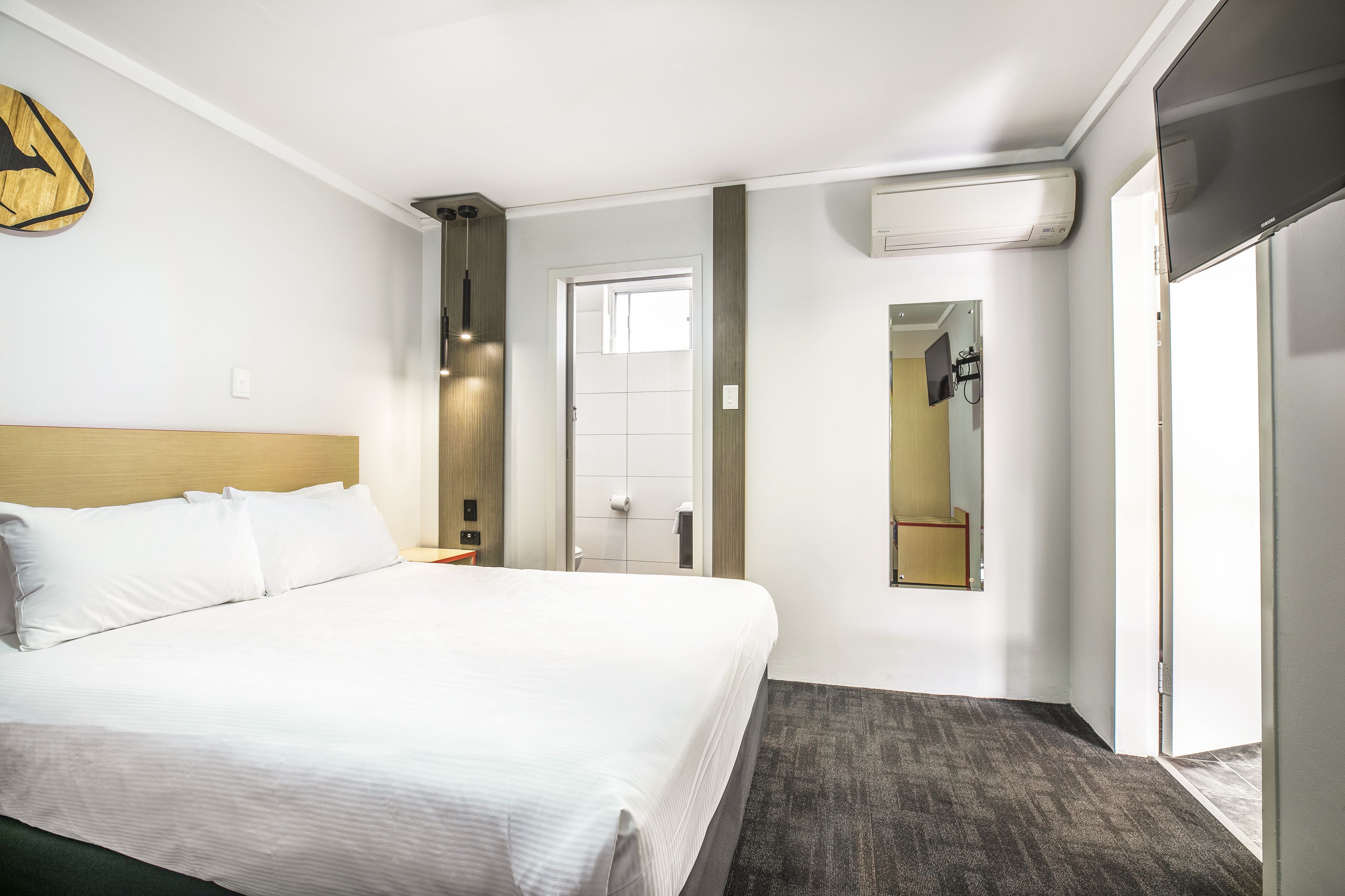 A hotel room with a large bed, a wooden headboard, a flat-screen TV, a mirror, and a door leading to a bathroom.
