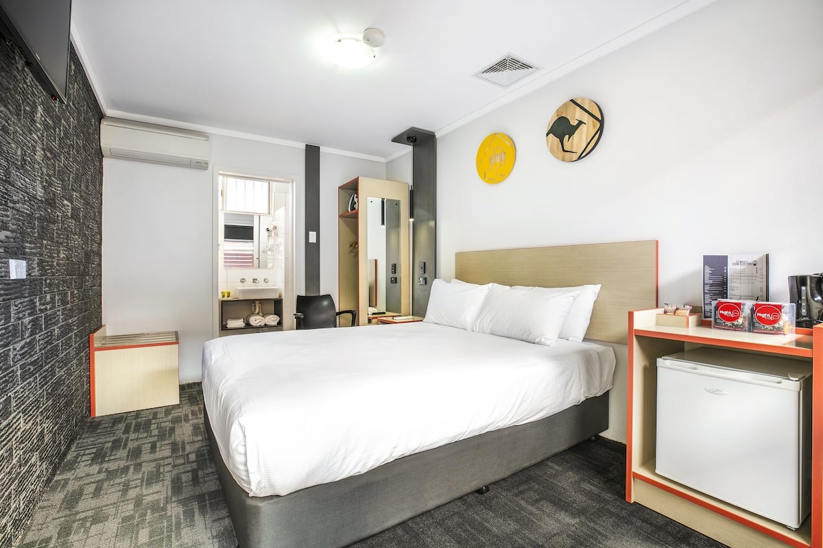 A hotel room with a bed, a small refrigerator, a desk with a coffee maker, a wardrobe, and a wall clock.