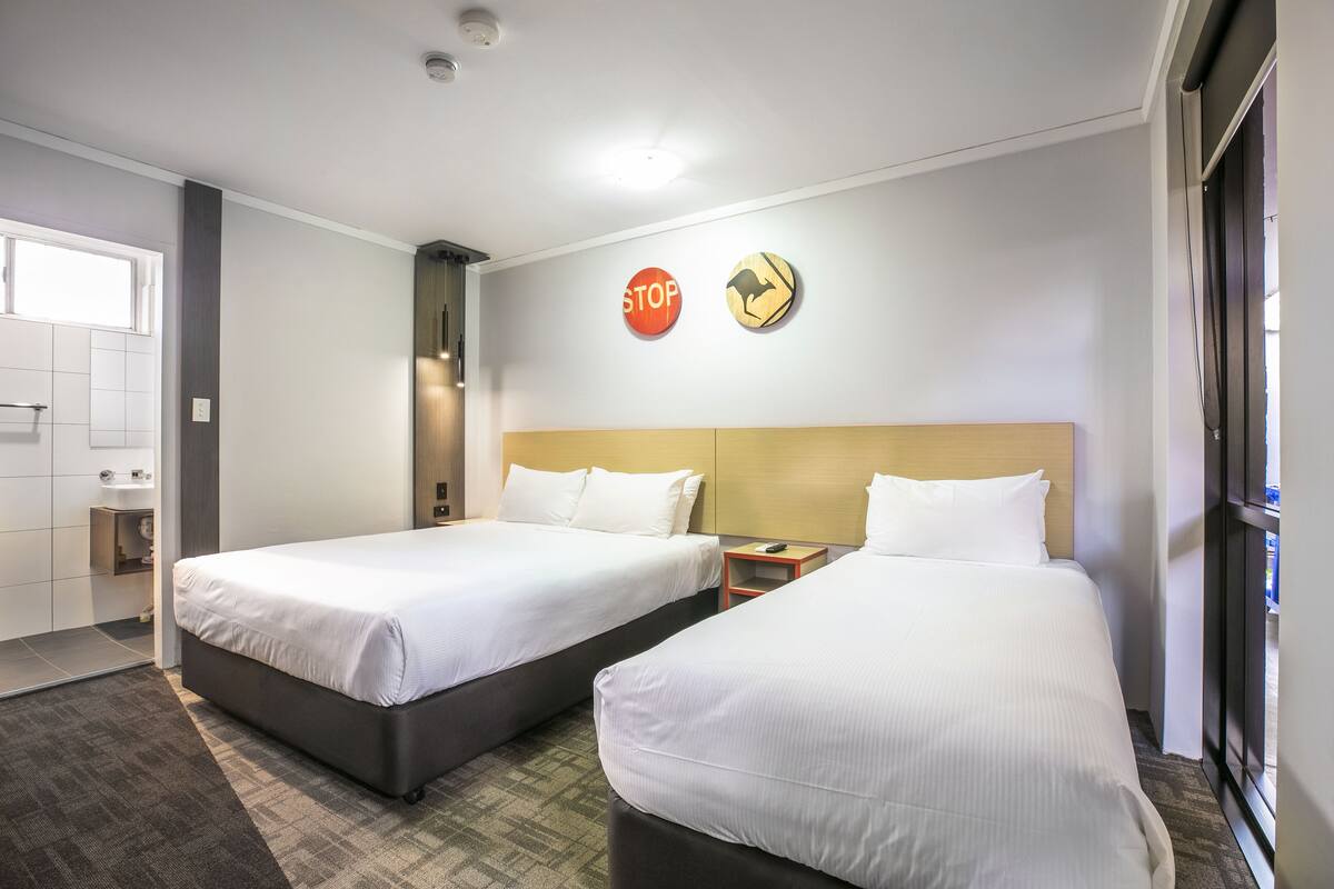 A hotel room with two beds, a small bedside table with a lamp, a wall-mounted clock, and a 'STOP' sign.