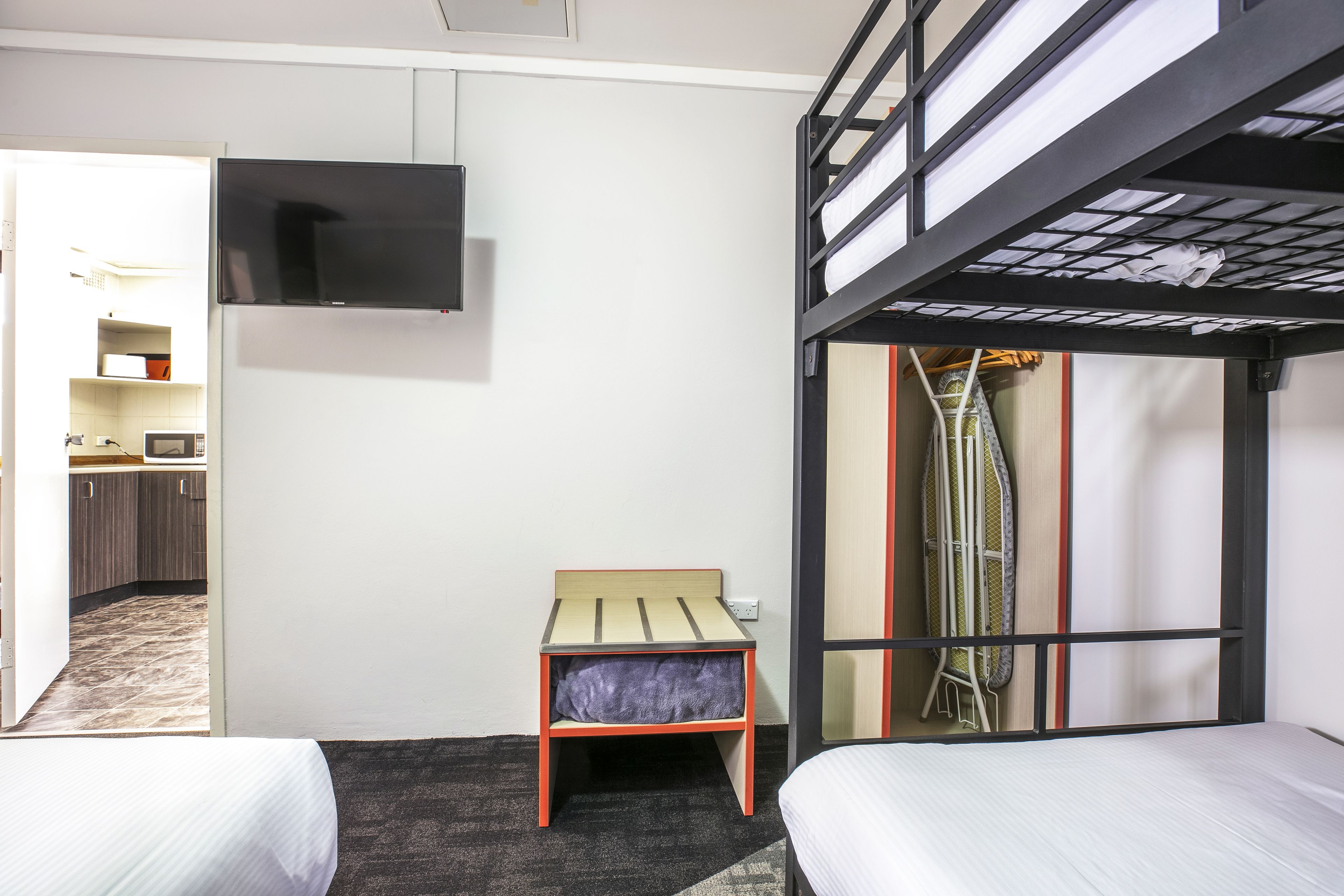 Habitación de hotel compacta con literas, una mesita pequeña, un televisor fijado a la pared y una kitchenette al fondo.