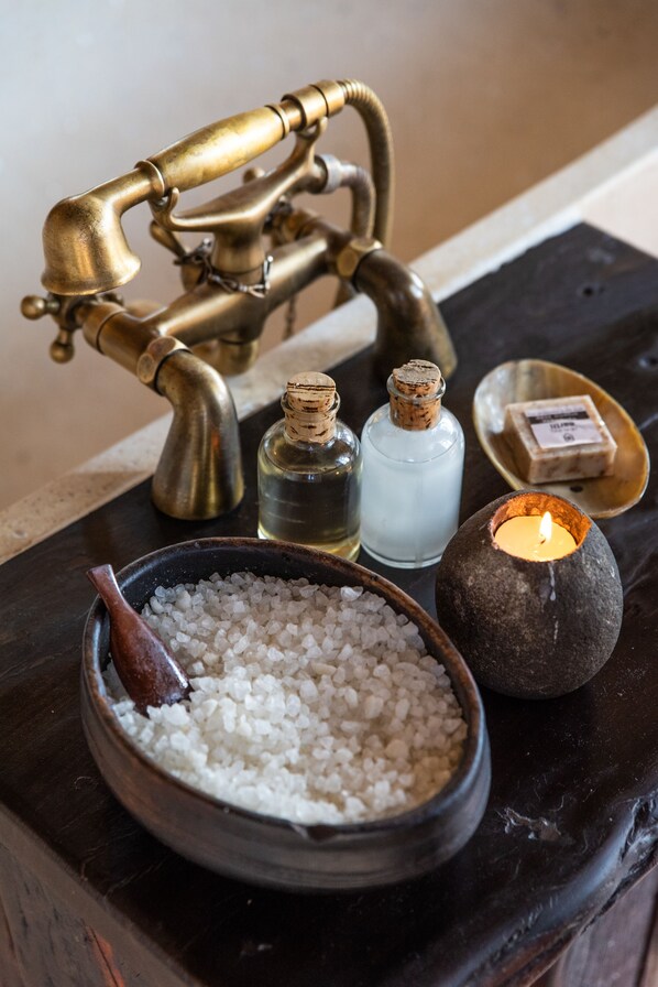 A bathroom vanity with a brass faucet, a bowl of white bath salts, a lit candle, and two bottles of liquid soap.
