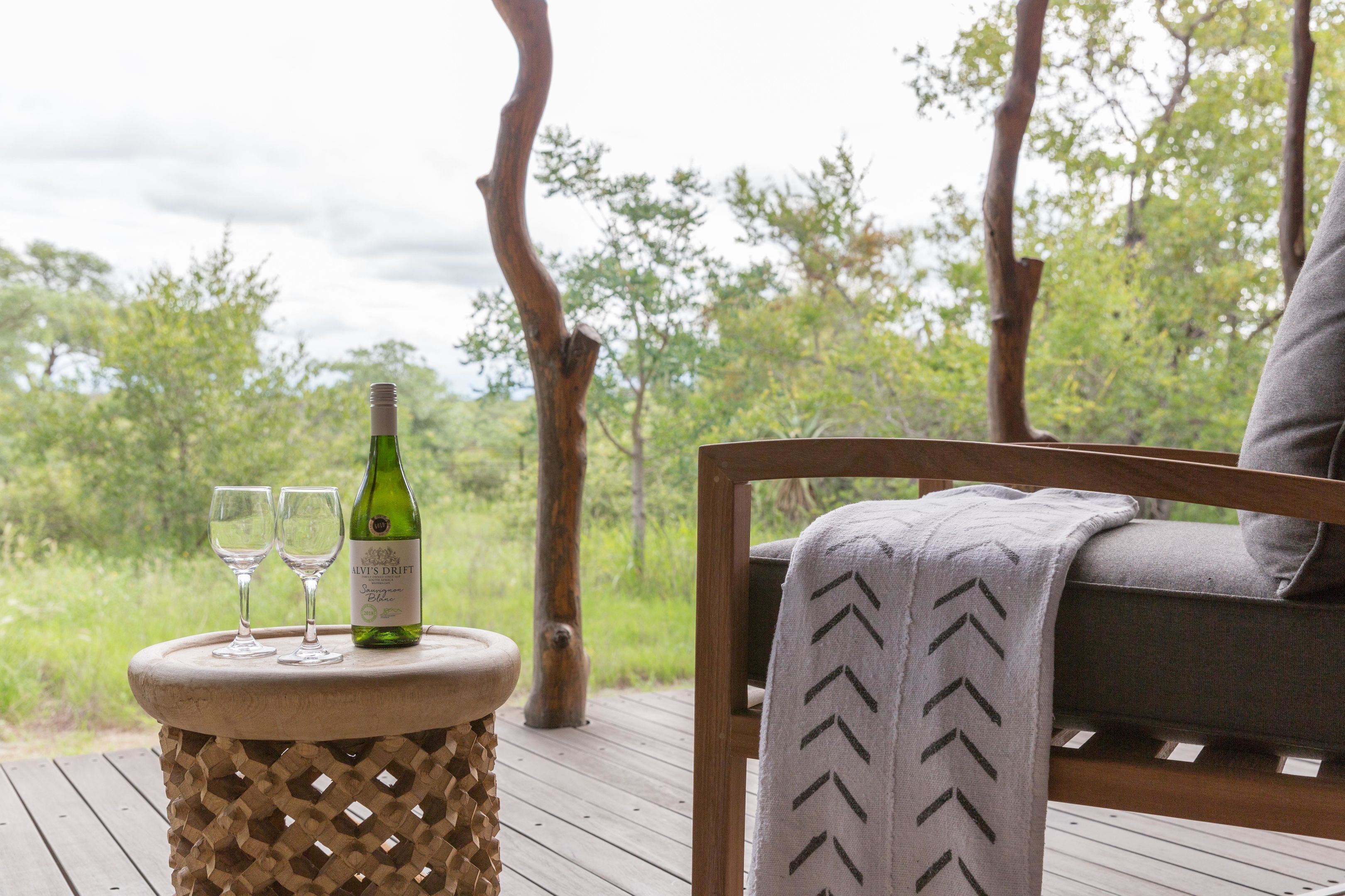 A wooden deck with a table holding a bottle and two glasses, a cushioned chair with a patterned throw, and a view of greenery.