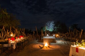 An outdoor dining area with tables set for a meal, a central fire pit, and a backdrop of trees and a night sky.