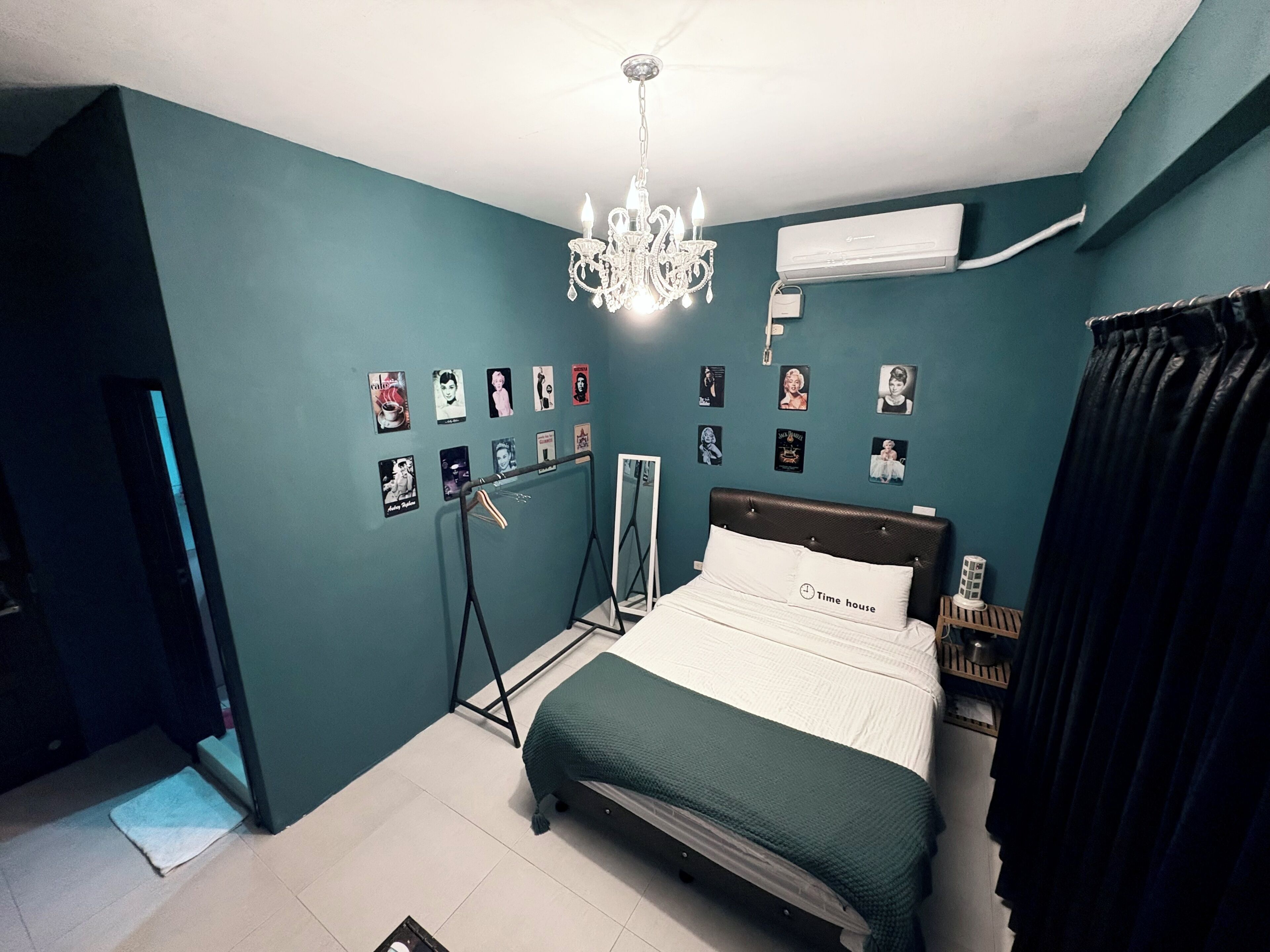 Une chambre avec des murs de couleur turquoise, un lit recouvert d’une couverture verte, un lustre, un climatiseur et divers affiches accrochées au mur.