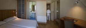 Habitación de hotel con cama, cabecera de madera, escritorio con lámpara y baño visible a través de una puerta abierta.
