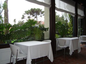 Un área de comedor al aire libre con mesas y sillas, rodeada de abundante vegetación y vistas a árboles y edificios.