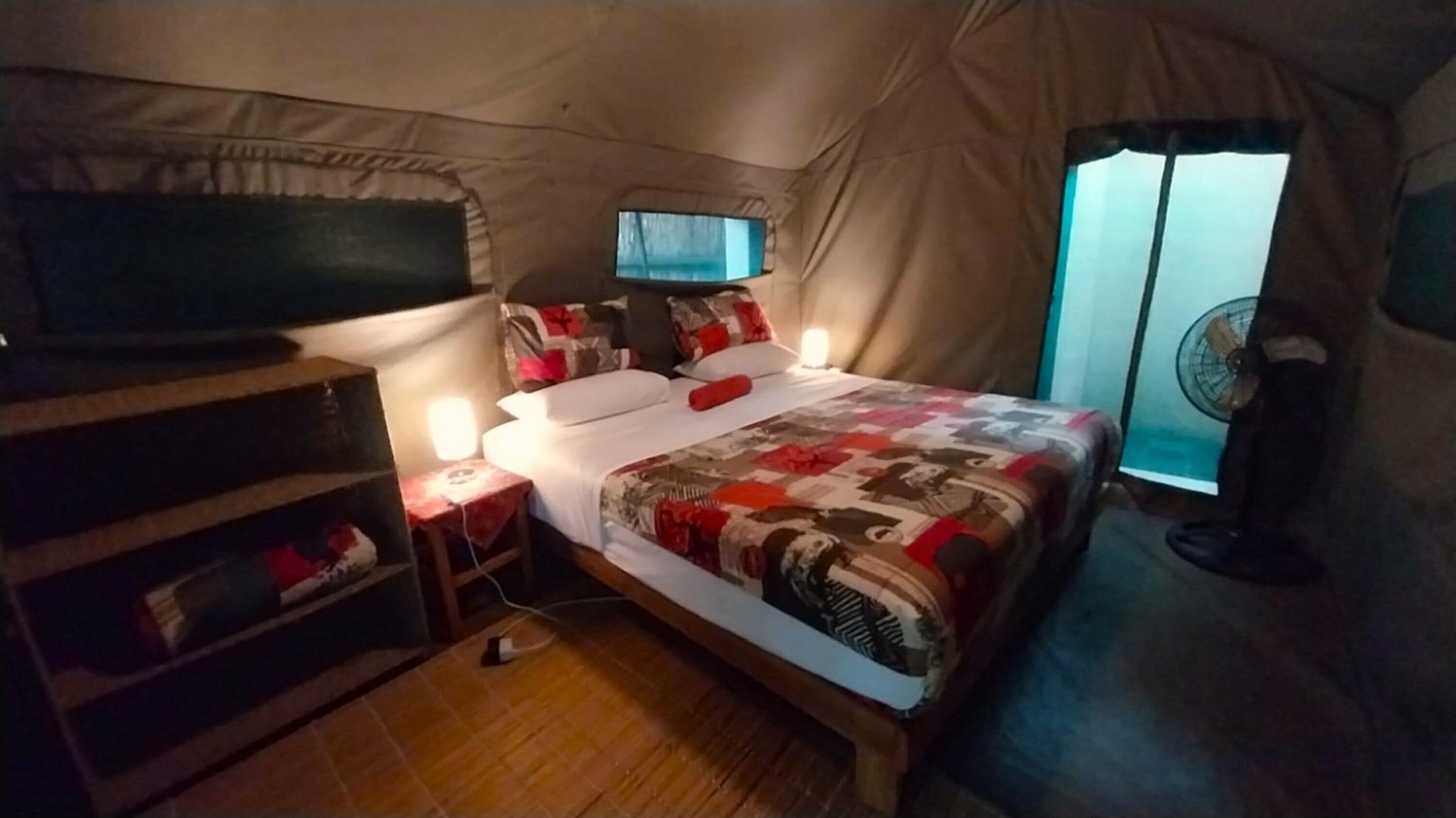 A tent-style room with a bed, a television, a fan, and a small table with a lamp.