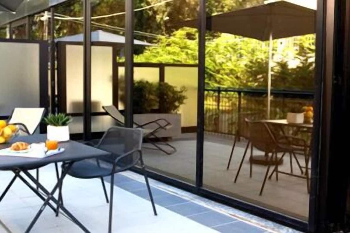 A modern outdoor dining area with a glass partition, black metal chairs, and a table set with food and drinks.