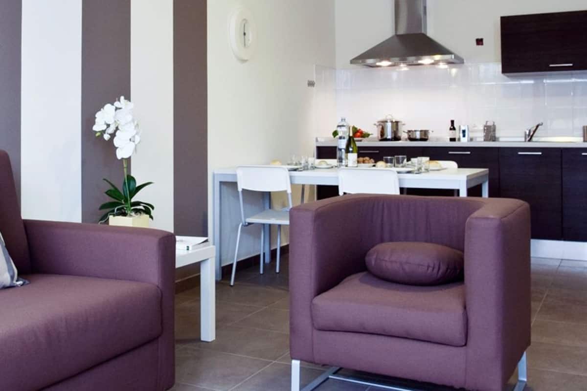 A modern living room with a sofa and armchair, a dining area with a table and chairs, and a kitchen with appliances and a TV.
